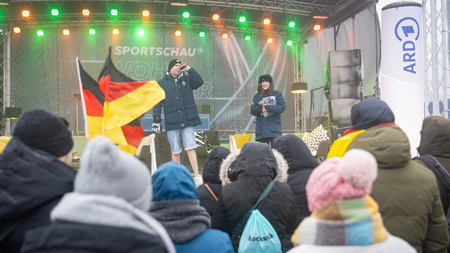 Das Bild zeigt eine Außenveranstaltung in Deutschland, bei der Zuschauer in Winterkleidung aufmerksam einer Person mit Mikrofon auf der 