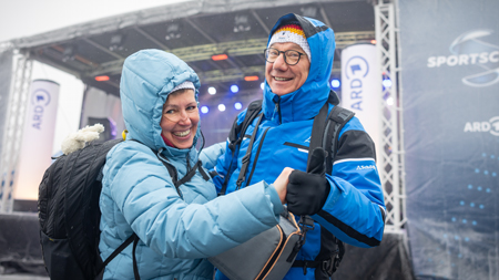 Das Bild zeigt zwei lächelnde Personen in blauen Winterjacken, die vor einer Bühne mit der Aufschrift 