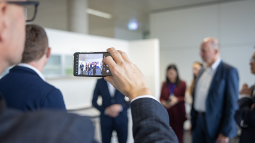 Person hält ein Smartphone hoch und fotografiert eine Gruppe von Geschäftsleuten, die unscharf im Hintergrund eines modernen Büros zu sehen sind.