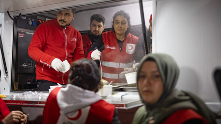Helfer des Roten Halbmonds verteilen Essen an Bedürftige aus einem mobilen Versorgungswagen heraus, während eine Frau im Vordergrund wartet.