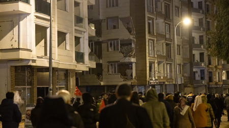 Menschenmengen auf der Straße bei Nacht, die ein beschädigtes Gebäude nach einem Erdbeben betrachten, mit sichtbaren Rissen und zerstörten Fassadenteilen.
