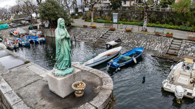Ein Luftbild einer Statue der Jungfrau Maria an einer Uferpromenade mit Blick auf eine kleine Hafenbucht, in der bunte Boote liegen, umgeben von Gebäuden und Bäumen, bei bedecktem Himmel.