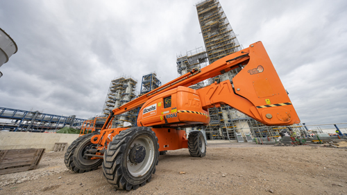 Die orangefarbene Hebebühne von Boels sticht vor dem industriellen Hintergrund hervor. Sie wirkt, als wäre sie für jede Herausforderung auf der Baustelle gewappnet, und der klar erkennbare Himmel deutet auf einen arbeitsreichen Tag hin.