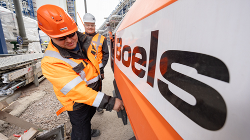 Zwei Bauarbeiter in reflektierender Schutzausrüstung und mit Helmen, die das Boels-Logo an einer Baustelle befestigen, vor dem industriellen Hintergrund des Chemieparks Leuna.