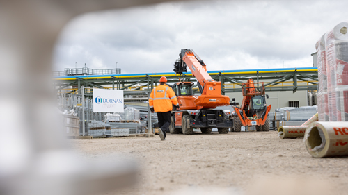 Ein Bauarbeiter in auffälliger orangefarbener Schutzkleidung geht an einem orangefarbenen Boels-Kran auf einer Baustelle vorbei, im Hintergrund sind industrielle Anlagen erkennbar.