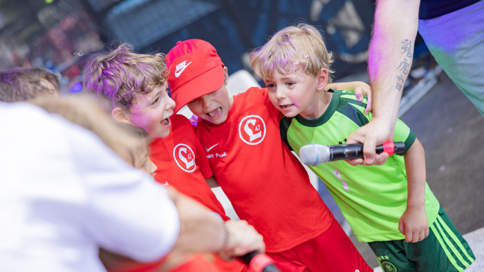 rei Kinder in roten und grünen Trikots singen fröhlich zusammen ins Mikrofon auf dem Fanfest des DFB-Pokalfinals in Berlin.