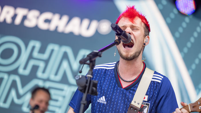 Ein Musiker mit roten Haaren und blauem Trikot singt leidenschaftlich ins Mikrofon auf der Bühne des Fanfests beim DFB-Pokalfinale in Berlin.