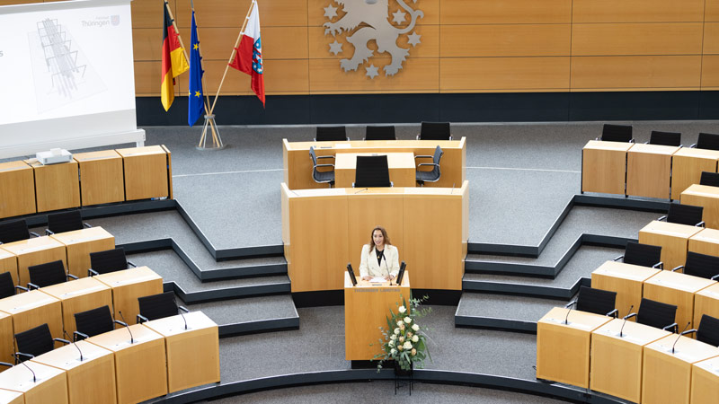 Eine Frau, Semiya Şimşek, steht an einem Rednerpult in einem modernen Landtagssaal. Im Hintergrund sind die Flaggen von Deutschland, Thüringen und der Europäischen Union sowie ein großes Emblem an der Holzwand zu sehen.