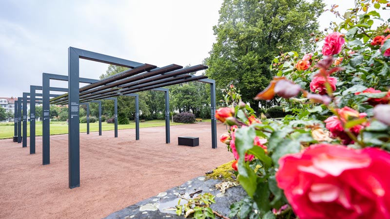 Eine moderne Außengedenkstätte aus Stahlrahmen in einem Park. Das Denkmal befindet sich auf einem roten Kiesweg mit einem Hintergrund aus üppigen grünen Bäumen. Im Vordergrund sind leuchtend rote Rosen zu sehen.