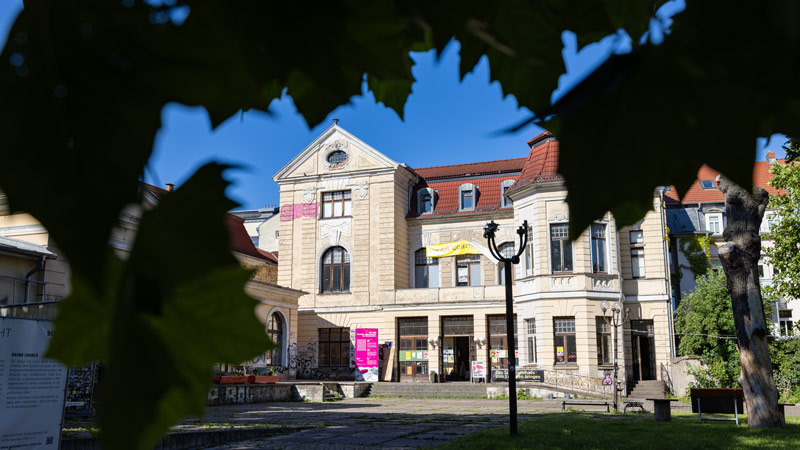 Außenansicht des Kulturquartier Schauspielhaus in Erfurt, das ein historisches Gebäude mit Bannern und Plakaten an der Fassade zeigt, sichtbar durch die Blätter der nahegelegenen Bäume.