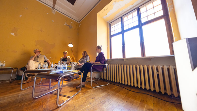 Eine Gruppendiskussion in einem Raum mit abblätternder gelber Farbe an den Wänden, vier Personen sitzen in einem Halbkreis in der Nähe eines großen Fensters.