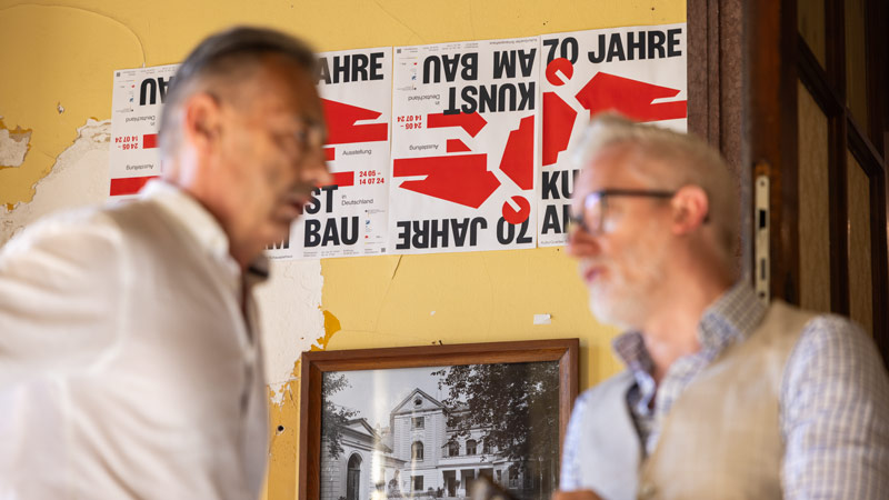 Zwei Männer im Gespräch vor einer Wand mit Plakaten, die '70 Jahre Kunst am Bau' lesen, in einem gelb gestrichenen Raum mit einem Schwarz-Weiß-Foto des Gebäudes im Hintergrund.