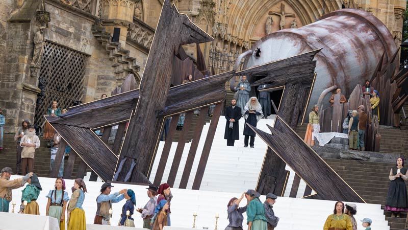 Ein Bühnenbild auf den Stufen eines historischen Gebäudes, vermutlich einer Kathedrale, bei einer Freilichtaufführung. In der Mitte befindet sich eine große, abstrakte Holzskulptur, die wie zersplitterte Balken aussieht. Mehrere Personen in historischen Kostümen stehen und sitzen auf der Bühne. Zwei zentrale Figuren in dunkler Kleidung stehen oben auf den Stufen, während unten mehrere Darsteller in bunten Gewändern versammelt sind. Im Hintergrund sind gotische Architekturdetails der Kathedrale sichtbar. Die Szene vermittelt eine dramatische und feierliche Atmosphäre.