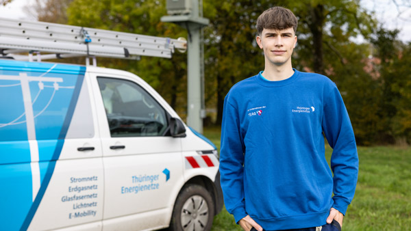 Ein junger Mann in einem blauen Sweatshirt mit der Aufschrift „Thüringer Energienetze“ steht vor einem Firmenfahrzeug. Im Hintergrund befinden sich eine grüne Wiese und ein Strommast.