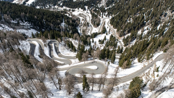 Eine beeindruckende Luftaufnahme von einer verschlungenen Bergstraße in der Schweiz, umgeben von verschneiten Wäldern und winterlicher Landschaft.