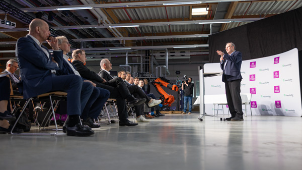 Ein Sprecher hält einen Vortrag vor einem Publikum in einer modernen Konferenzhalle in Arnstadt. Im Hintergrund sind eine Leinwand und industrielle Maschinen sichtbar, die auf das Thema der Veranstaltung hinweisen.