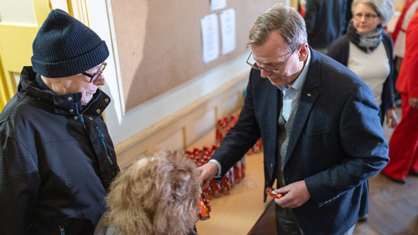 Ein Mann in einem Anzug überreicht einer älteren Frau ein kleines Geschenk, während ein weiterer älterer Mann daneben steht. Im Hintergrund sind weitere Geschenke zu sehen, die für die Verteilung vorbereitet wurden.