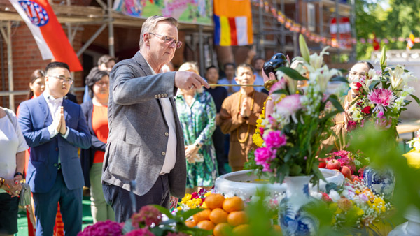 Ein Mann in einem Anzug steht vor einem blumengeschmückten Altar und nimmt an einem traditionellen Ritual teil. Im Hintergrund stehen weitere Personen, die respektvoll zuschauen.