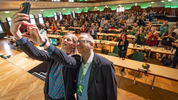 Eine Frau und ein Mann lächeln in die Kamera eines Smartphones, während sie ein Selfie aufnehmen. Im Hintergrund ist ein gut besuchter Saal mit applaudierenden Menschen zu sehen, die eine Veranstaltung feiern.