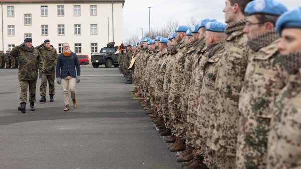 Eine Gruppe von Soldaten in Uniformen mit blauen Baretten steht in einer Reihe, während ein Mann mit einem Offizier die Formation entlanggeht. Im Hintergrund sind ein Gebäude und Militärfahrzeuge sichtbar.