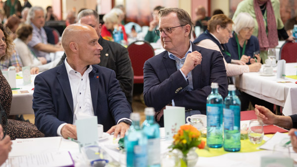 wei Männer in Anzügen sitzen in einem Konferenzsaal an einem Tisch, umgeben von anderen Teilnehmern. Wasserflaschen und Blumenarrangements befinden sich auf den Tischen. Die Szene zeigt ein politisches oder geschäftliches Gespräch.