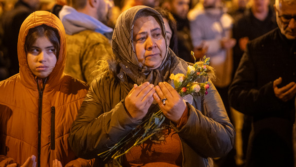 Eine Frau hält Blumen in den Händen und steht mit ernster, nachdenklicher Miene bei einer nächtlichen Gedenkveranstaltung. Neben ihr ein Kind in einer orangen Jacke. Im Hintergrund sind Menschen in stiller Anteilnahme zu sehen.