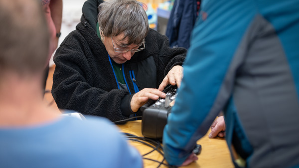 Eine ältere Person arbeitet konzentriert an einem technischen Gerät während eines Repair Cafés oder einer ähnlichen Veranstaltung. Umgeben von anderen Teilnehmern herrscht eine engagierte und gemeinschaftliche Atmosphäre.