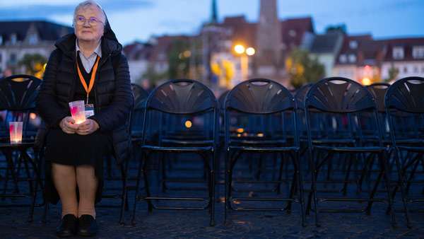 Eine ältere Frau sitzt mit einer Kerze in der Hand auf einer der vielen leeren Stuhlreihen auf einem Platz in Erfurt. Die Szene spielt in der Dämmerung, mit stimmungsvoller Beleuchtung im Hintergrund.