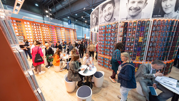 Eine lebhafte Szene auf einer Messe in Leipzig: Menschen schlendern durch einen farbenfrohen Standbereich, einige sitzen an Tischen und führen Gespräche, während im Hintergrund Bücher und Bilder die Stände schmücken.