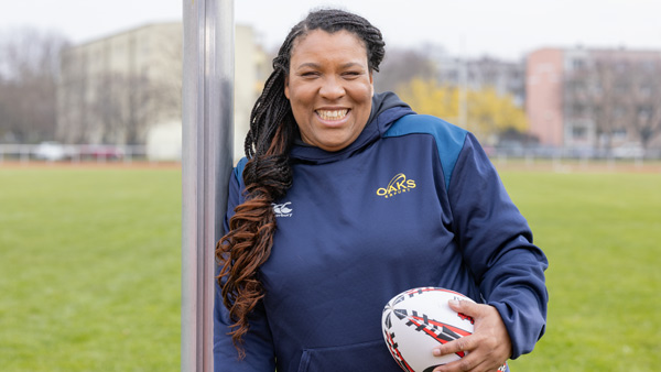 Eine Frau in sportlicher Kleidung, die ein Rugby-Ball hält, lächelt in die Kamera. Im Hintergrund ist ein grünes Sportfeld zu sehen, das auf eine Trainings- oder Wettkampfsituation in Erfurt oder Thüringen hinweist.