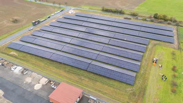 Eine Drohnenaufnahme eines großflächigen Solarparks mit mehreren Reihen von Solarpaneelen, die auf einer grünen Fläche installiert sind. Am Rand sind Gebäude und Fahrzeuge zu erkennen.