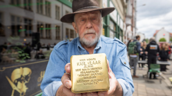 Ein älterer Mann mit Hut hält einen Stolperstein mit der Aufschrift „Hier wohnte Karl Klaar, Jahrgang 1890, seit 1934 in mehreren Heilanstalten, ‚verlegt‘ 28.10.1940 Bernburg, ermordet 28.11.1940 ‚Aktion T4‘“. Im Hintergrund ist eine Straße mit Passanten zu sehen.