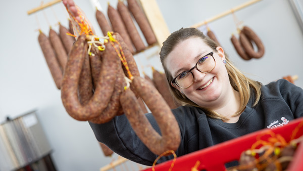 Eine Frau in einer Wursterei präsentiert lächelnd mehrere frische Würste. Im Hintergrund hängen weitere Wurstwaren in einer traditionellen Produktionsumgebung.