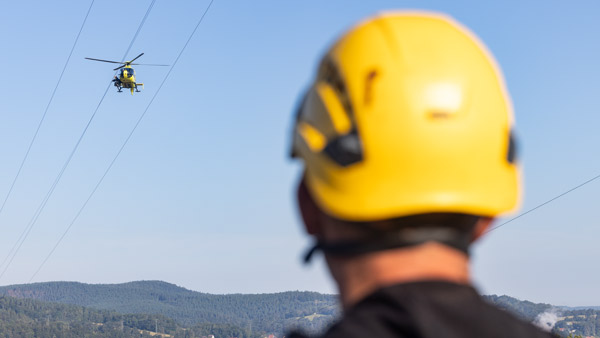 Ein Mitarbeiter mit gelbem Schutzhelm beobachtet einen Hubschrauber, der in der Ferne über Stromleitungen schwebt. Im Hintergrund sind bewaldete Hügel unter klarem Himmel zu sehen.