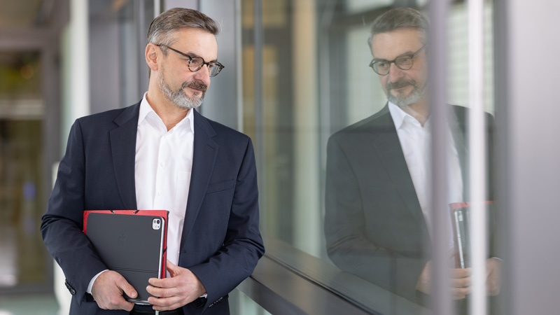 Geschäftsmann mit Brille im Anzug hält ein Tablet und blickt nachdenklich durch ein Fenster, in dem sich sein Spiegelbild zeigt.