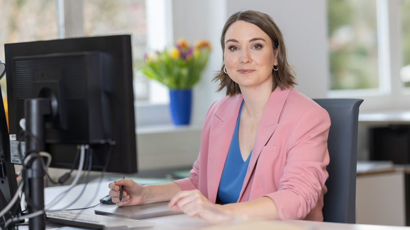 Geschäftsfrau in einem rosafarbenen Blazer sitzt an einem Schreibtisch vor einem Computerbildschirm und hält einen Stift, im Hintergrund steht ein bunter Tulpenstrauß.