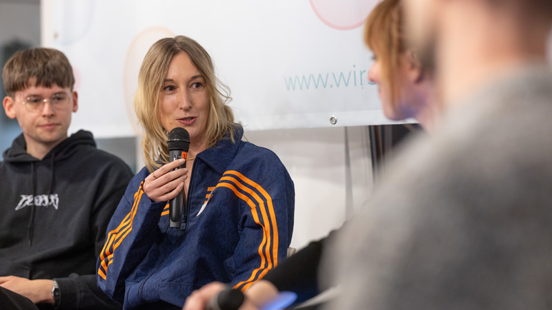 Eine Frau in einer blauen Trainingsjacke mit orangenen Streifen spricht in ein Mikrofon, während zwei weitere Personen aufmerksam zuhören.