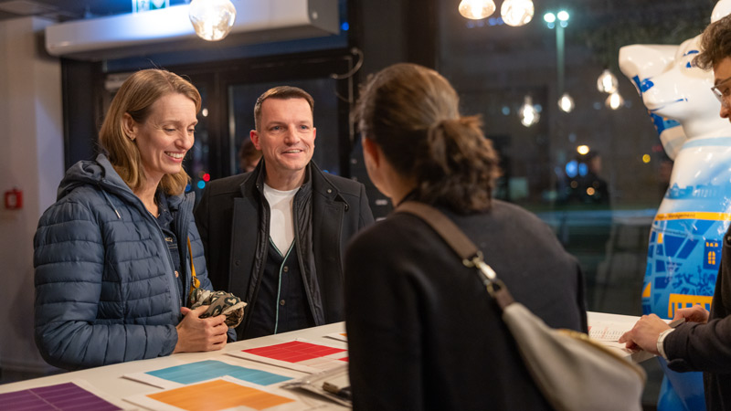 Zwei lächelnde Personen, ein Mann und eine Frau, stehen an einem Empfangstresen und sprechen mit einer anderen Frau in einer warm beleuchteten Umgebung.