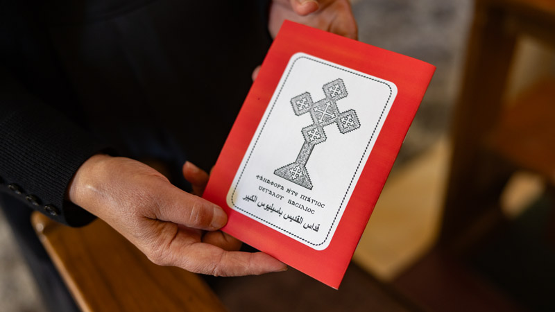 Hände halten ein rotes Kirchenheft mit einem koptischen Kreuz und arabisch-griechischer Schrift auf dem Umschlag.