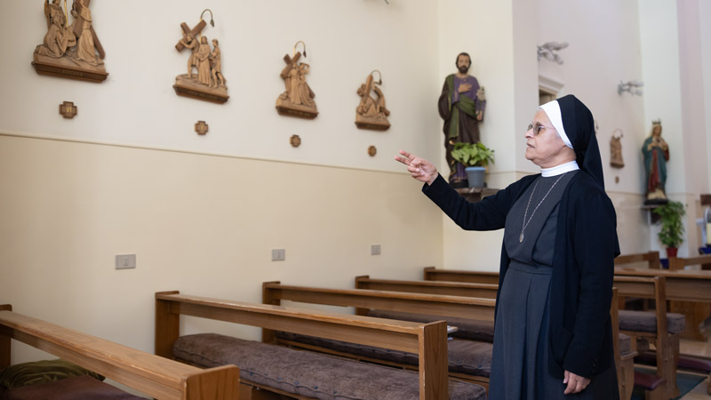 Nonne zeigt auf Wandreliefs mit Kreuzweg-Szenen in einer Kapelle mit Holzsitzreihen und Heiligenfiguren.