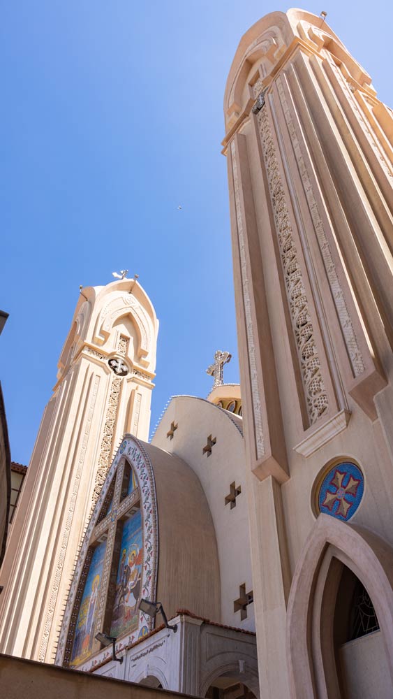Hohe Türme einer koptisch-orthodoxen Kirche mit reich verzierten Fassaden und bunten Wandmosaiken vor blauem Himmel.