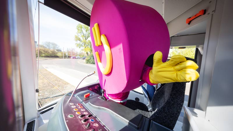 REWE Bonus-Maskottchen sitzt am Steuerpult der Straßenbahn und winkt aus dem Fahrerbereich.
