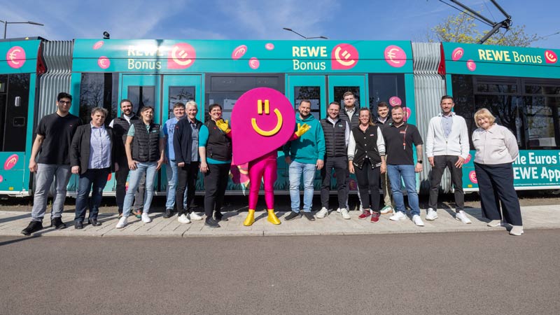 Team von Menschen posiert mit REWE Bonus-Maskottchen vor gebrandeter Straßenbahn auf dem Gehweg.