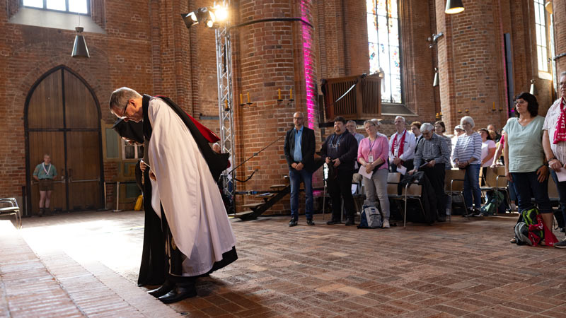 Älterer Geistlicher verbeugt sich in einer Kirche, während Menschen in den Bänken respektvoll stehen.