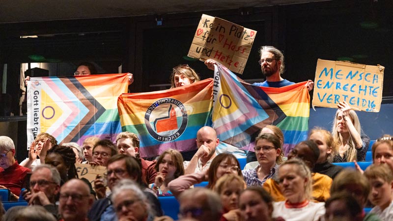 Teilnehmende in einer Halle halten Regenbogenfahnen und Plakate mit Botschaften gegen Hass und für Menschenrechte hoch.