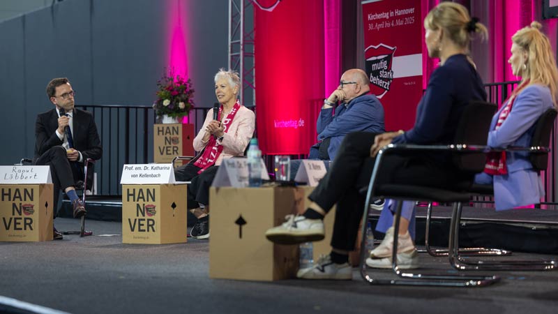 Fünf Personen diskutieren auf einer Bühne mit roten Bannern, Teil des Kirchentags in Hannover 2025.