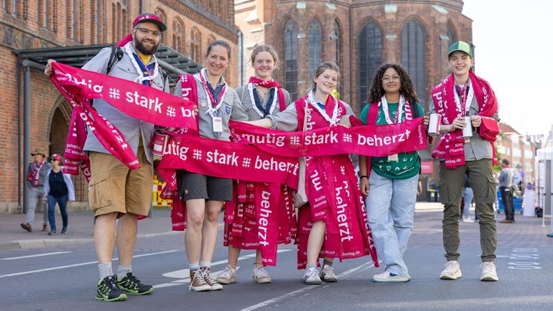 Sechs junge Menschen mit roten Kirchentag-Schals stehen lächelnd vor einer Kirche und halten Banner mit „mutig # stark # beherzt“.