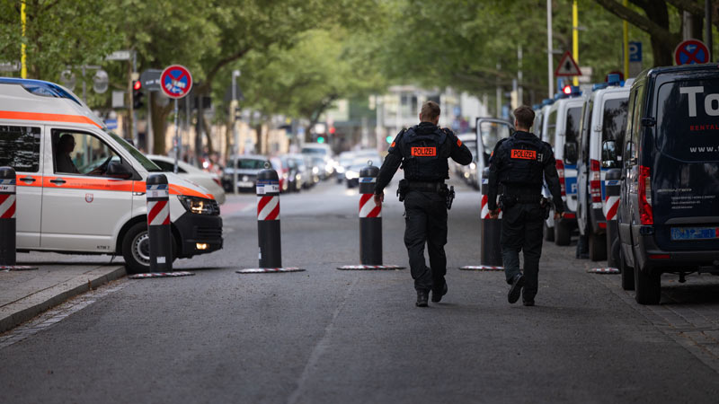 Zwei Polizisten gehen zwischen Polizeiwagen durch eine gesperrte Straße mit Absperrpfosten.