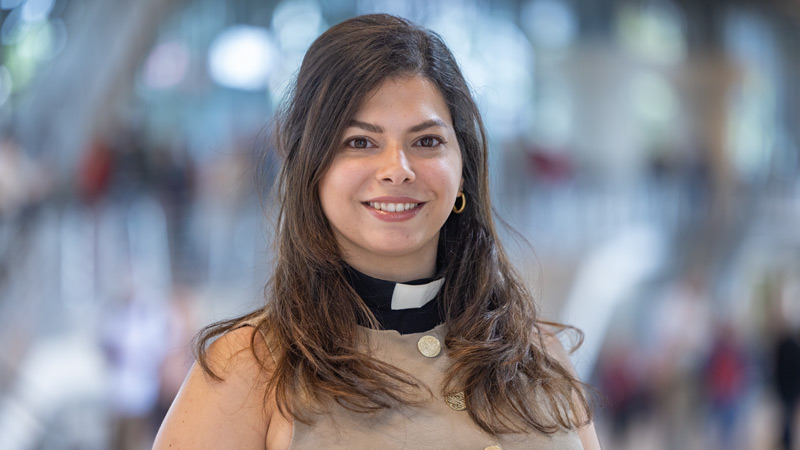 Junge Frau mit Priesterkragen und langen Haaren lächelt in einem hellen Gebäude in die Kamera.