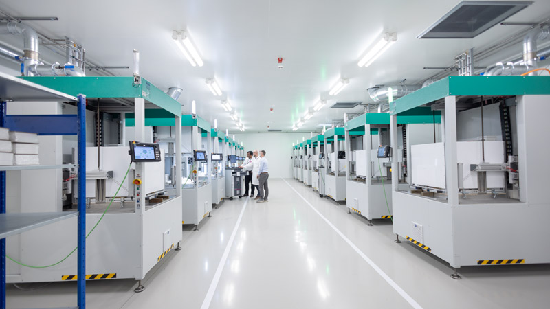 Moderne Fertigungshalle mit zwei Reihen automatisierter Produktionsmaschinen und drei Technikern im Hintergrund.